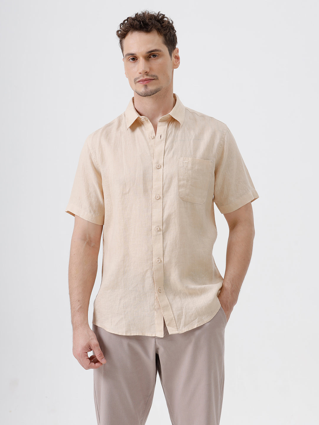 A man wearing a light beige short-sleeve linen button-up shirt and taupe trousers, standing against a plain white studio background.