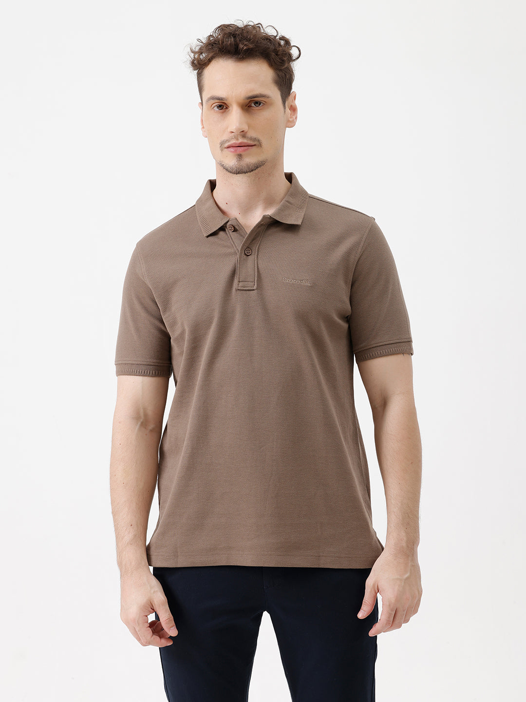 A man wearing a brown short-sleeve polo shirt with dark navy pants, standing against a plain light gray studio background.