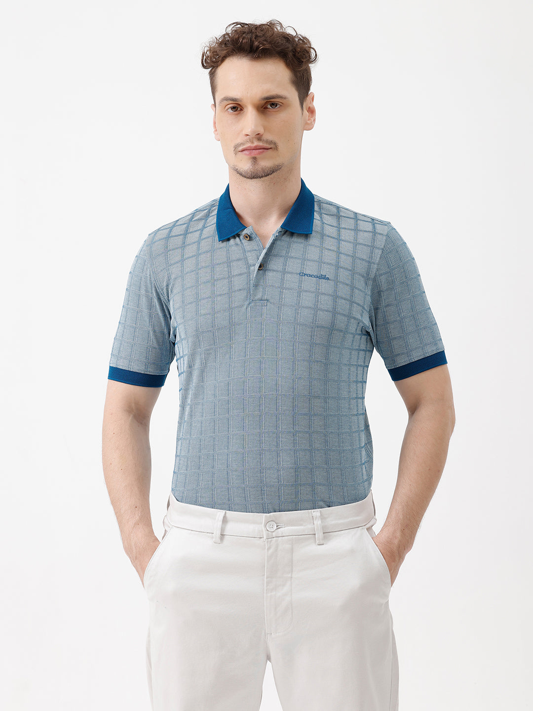 A man wearing a light blue and white checkered polo shirt with a dark blue collar and sleeve cuffs, paired with white trousers, standing against a plain white background.