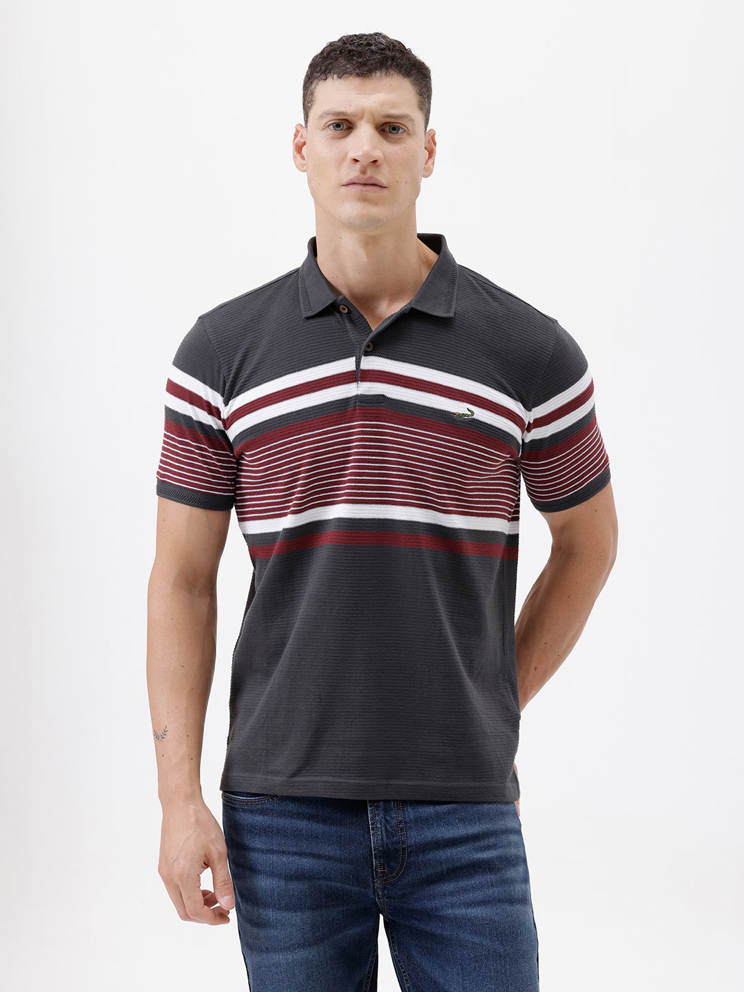A man wearing a dark gray Lacoste polo shirt with horizontal red and white stripes across the chest and sleeves, paired with blue jeans, against a plain light gray background.