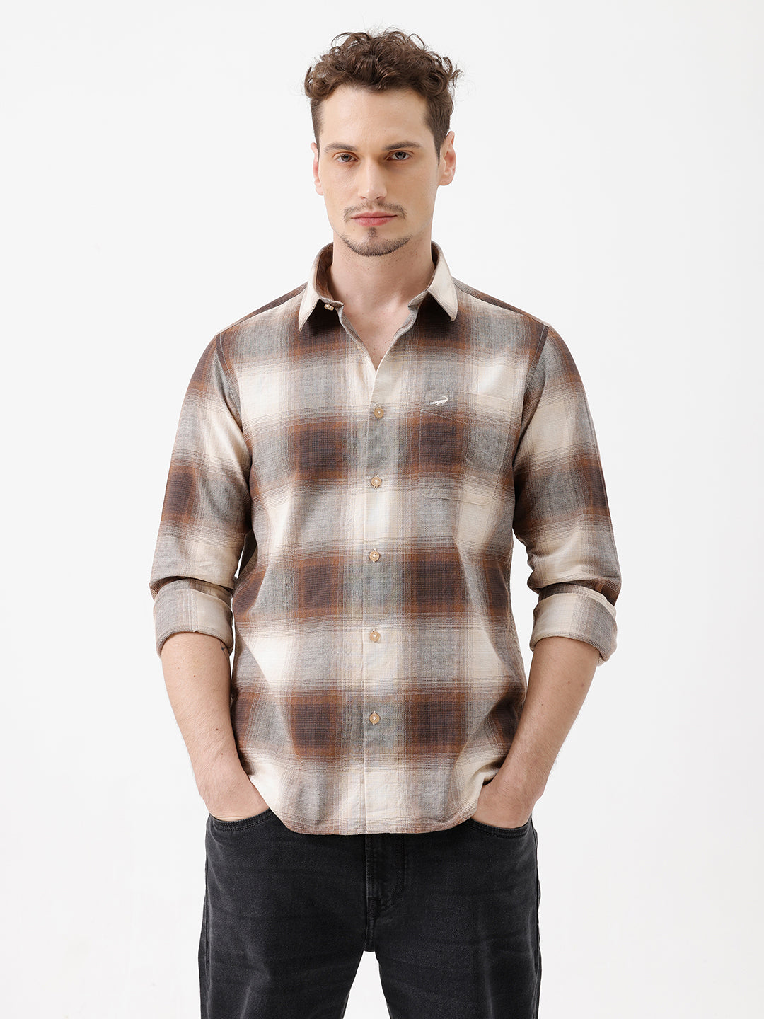 A man wearing a long-sleeve button-up plaid shirt in brown, beige, and gray tones, paired with dark jeans, standing against a plain white studio background.