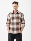 A man wearing a long-sleeve button-up plaid shirt in brown, beige, and gray tones, paired with dark jeans, standing against a plain white studio background.