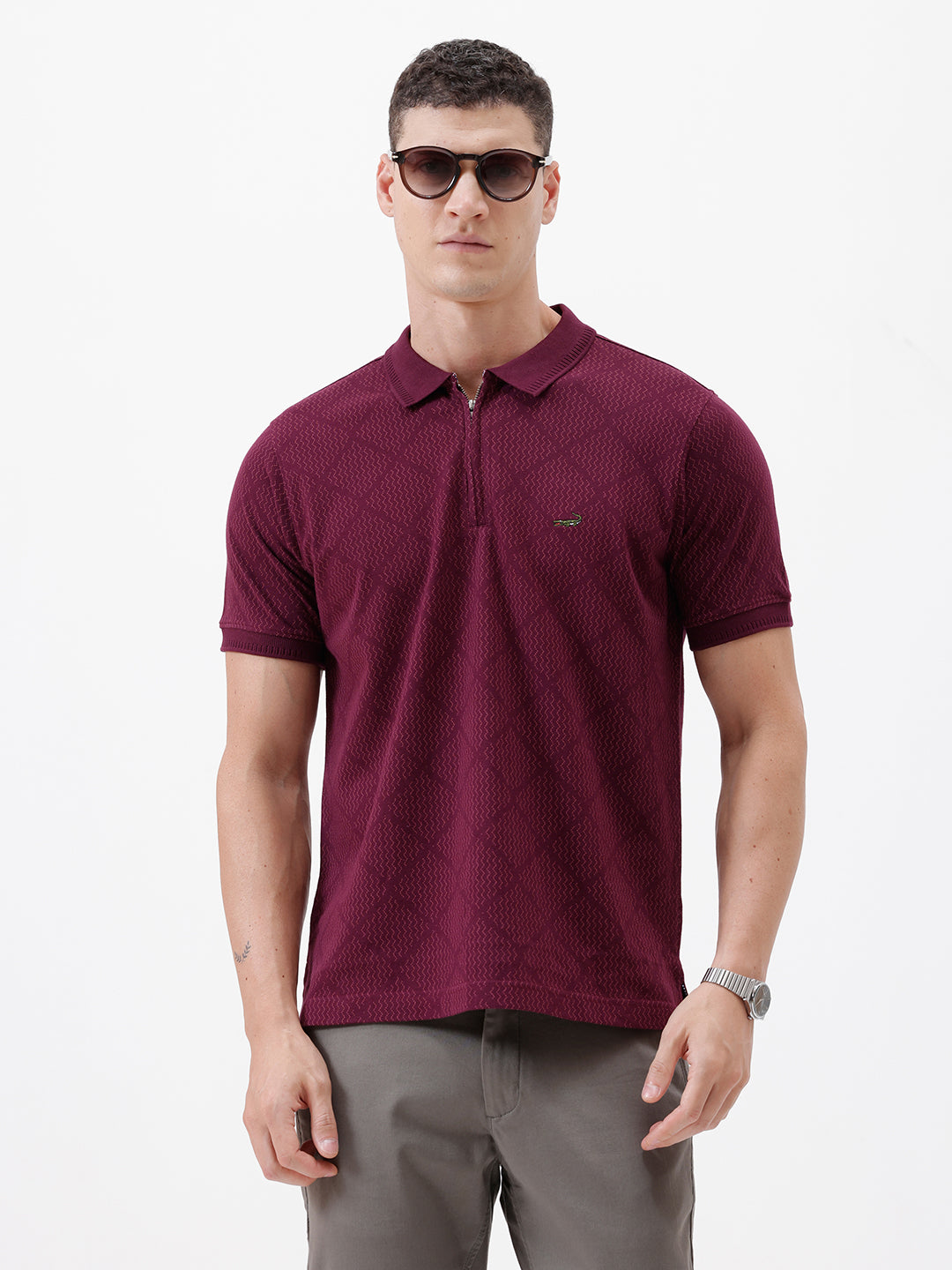 A man wearing a short-sleeve polo shirt in deep burgundy with a subtle textured geometric pattern, styled with gray trousers, sunglasses, and a watch against a plain white background.