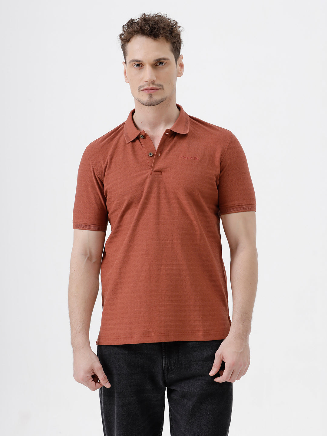 A man wearing a rust-orange short-sleeve polo shirt with a subtle horizontal stripe pattern, paired with dark jeans, standing against a plain white studio background.