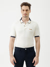 A man wearing a short-sleeve polo shirt in off-white with navy blue and gold trim on the collar and sleeve cuffs, paired with dark navy trousers, against a plain light gray background.