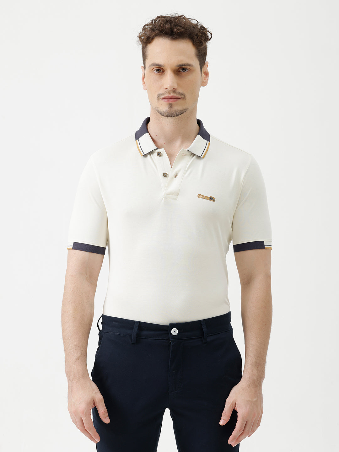 A man wearing a short-sleeve polo shirt in off-white with navy blue and gold trim on the collar and sleeve cuffs, paired with dark navy trousers, against a plain light gray background.