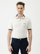 A man wearing a short-sleeve polo shirt in off-white with navy blue and gold trim on the collar and sleeve cuffs, paired with dark navy trousers, against a plain light gray background.