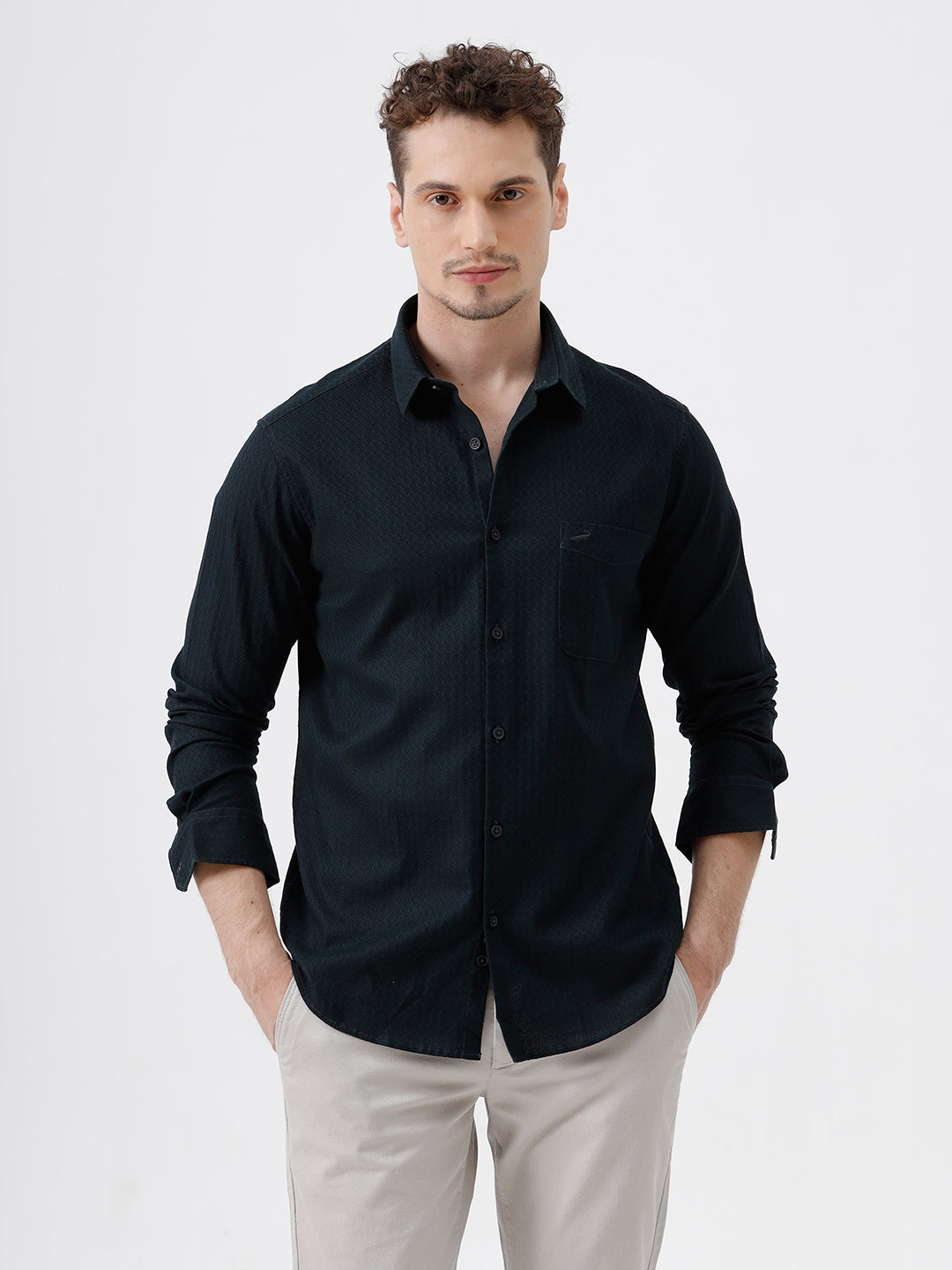 A man wearing a dark navy long-sleeve button-up shirt with a subtle textured pattern and light beige trousers, standing against a plain light gray background.
