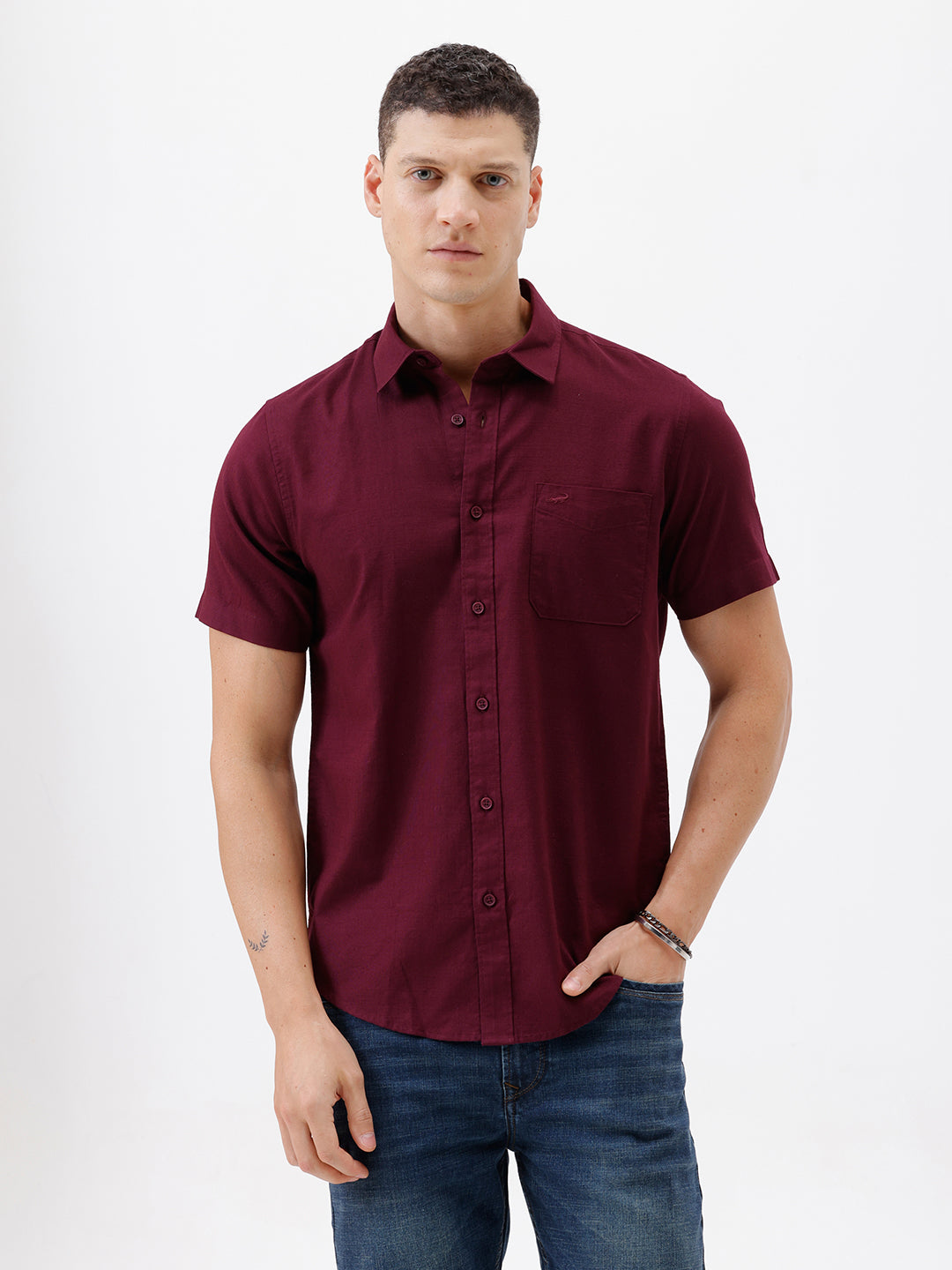A man wearing a short-sleeve button-up shirt in maroon with a chest pocket, paired with blue jeans, standing against a plain white studio background.