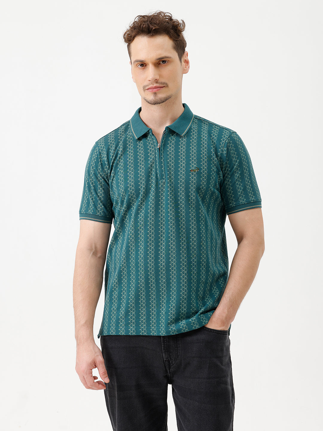 A man wearing a teal green short-sleeve polo shirt with a white geometric pattern, paired with black jeans, standing against a plain white studio background.