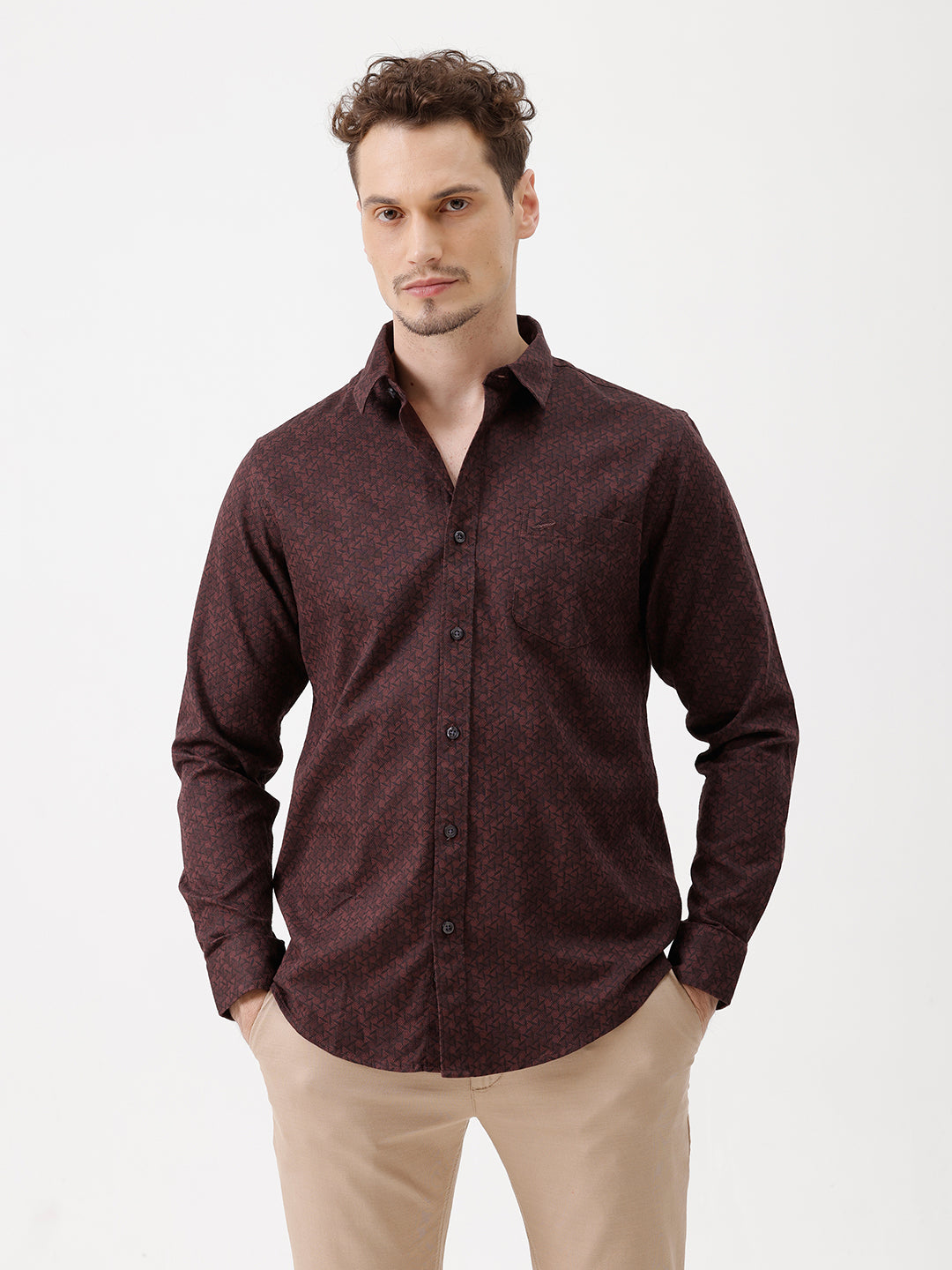 A man wearing a dark burgundy long-sleeve button-up shirt with a subtle geometric pattern, paired with beige trousers, standing against a plain white studio background.