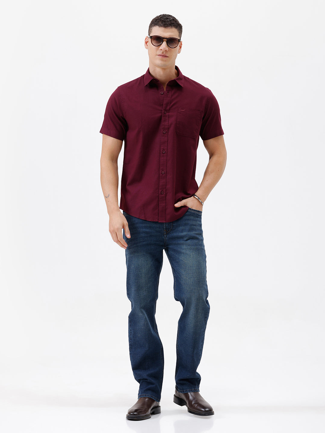 A man wearing a short-sleeve button-up shirt in maroon with a chest pocket, paired with blue jeans, standing against a plain white studio background.