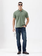 A man wearing a short-sleeve polo shirt in sage green, paired with dark blue jeans, standing against a plain light gray background.