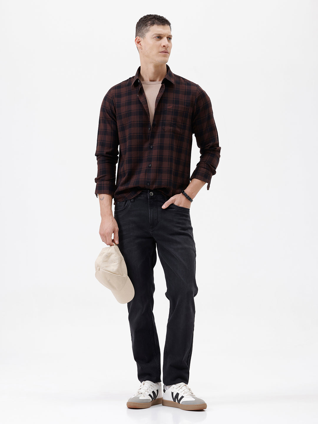 A man wearing a dark brown and black plaid long-sleeve button-up shirt, paired with dark pants, standing against a plain light gray background.