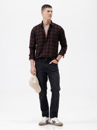A man wearing a dark brown and black plaid long-sleeve button-up shirt, paired with dark pants, standing against a plain light gray background.