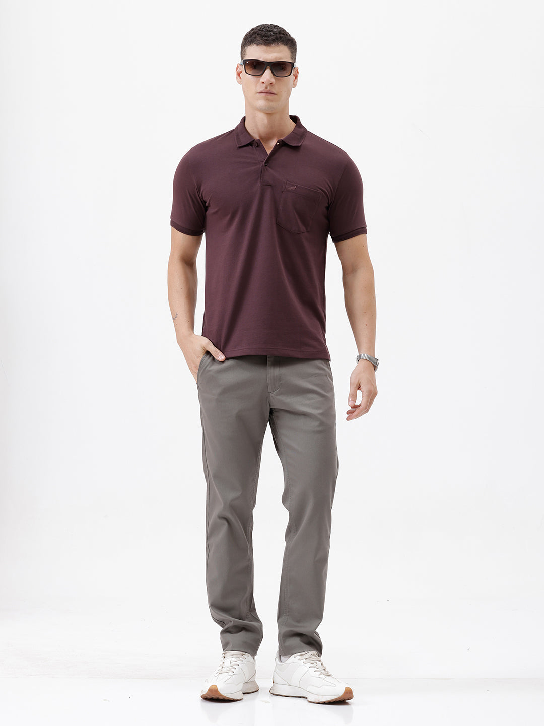 A man wearing a short-sleeve polo shirt in dark burgundy with a chest pocket, paired with gray trousers, standing against a plain light gray background.