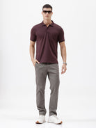 A man wearing a short-sleeve polo shirt in dark burgundy with a chest pocket, paired with gray trousers, standing against a plain light gray background.