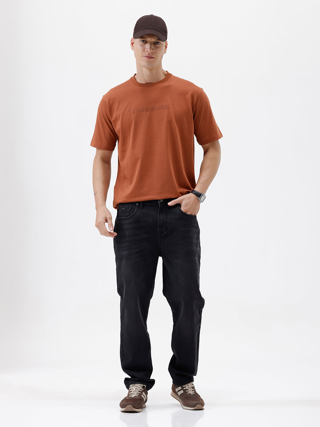A person wearing black slim-fit jeans, paired with a rust-orange t-shirt and brown sneakers, shown from the waist down against a plain white studio background.