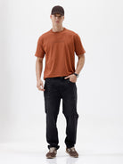 A person wearing black slim-fit jeans, paired with a rust-orange t-shirt and brown sneakers, shown from the waist down against a plain white studio background.