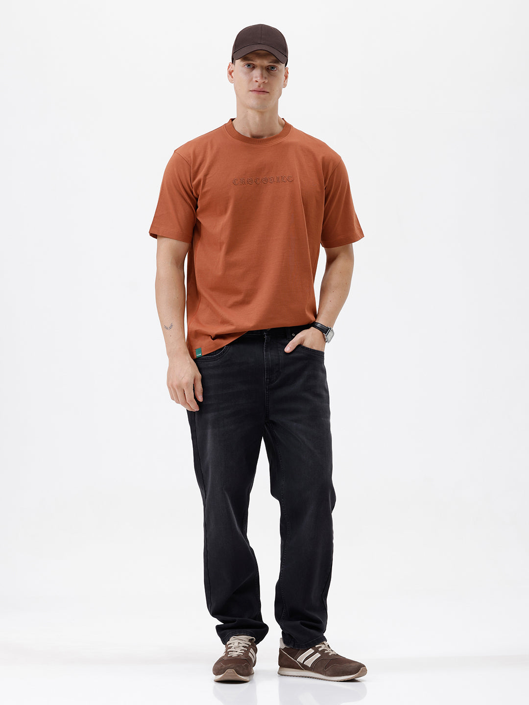 A man wearing a rust-orange crewneck t-shirt with a subtle tonal 'ELECTRIC' logo across the chest, paired with black pants, photographed against a clean white studio background.