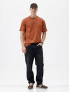 A man wearing a rust-orange crewneck t-shirt with a subtle tonal 'ELECTRIC' logo across the chest, paired with black pants, photographed against a clean white studio background.