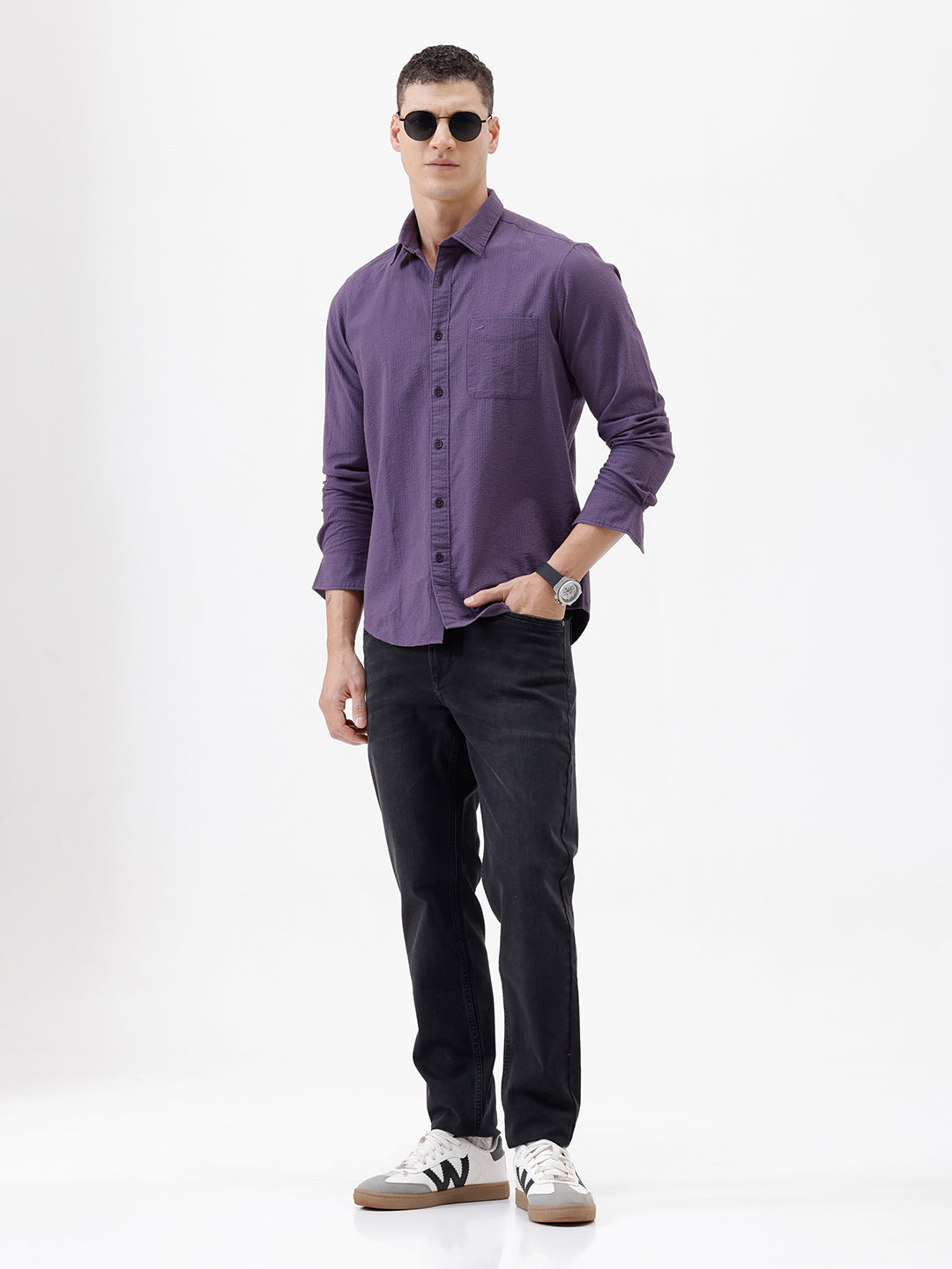 A man wearing a long-sleeve button-down shirt in a muted purple color, paired with black pants, standing against a plain white studio background.