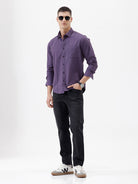 A man wearing a long-sleeve button-down shirt in a muted purple color, paired with black pants, standing against a plain white studio background.
