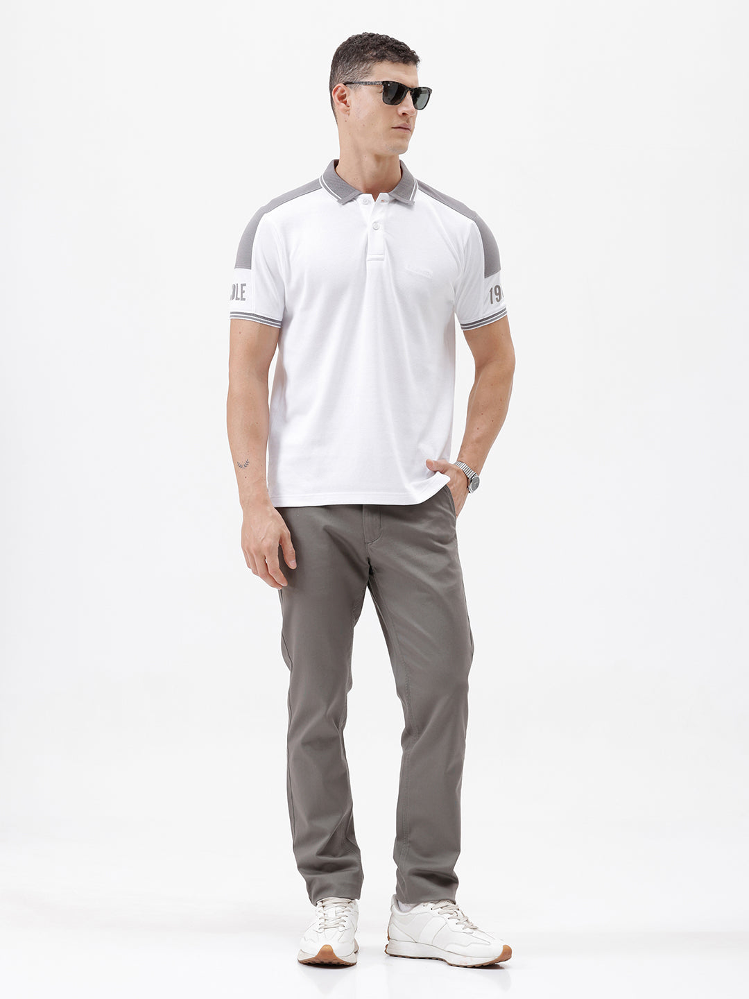 A man modeling a white short-sleeve polo shirt with gray shoulder panels and gray striped trim on the sleeves, paired with gray trousers and black sunglasses, against a plain white background.