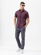 A man modeling a short-sleeve button-up shirt in a deep plum purple color, paired with gray trousers, against a plain white studio background.