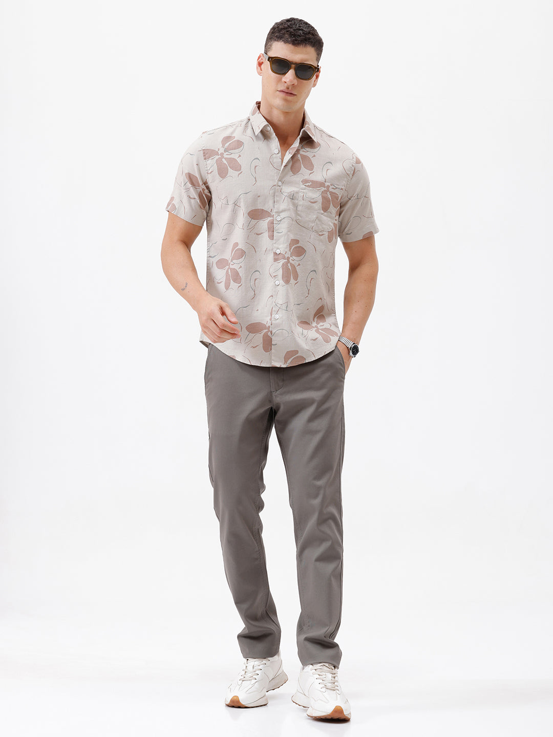 A man wearing a short-sleeve button-up shirt with a beige and brown floral pattern, paired with gray trousers, standing against a plain white background.