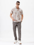 A man wearing a short-sleeve button-up shirt with a beige and brown floral pattern, paired with gray trousers, standing against a plain white background.