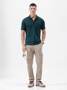 A man wearing a dark teal short-sleeve polo shirt with a chest pocket, paired with beige cargo pants, standing against a plain white studio background.