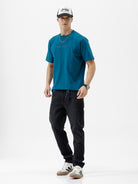 A man wearing a teal blue short-sleeve t-shirt with a subtle tonal 'GREATNESS' text graphic across the chest, paired with black pants, photographed against a clean white studio background.