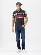 A man wearing a dark gray Lacoste polo shirt with horizontal red and white stripes across the chest and sleeves, paired with blue jeans, against a plain light gray background.