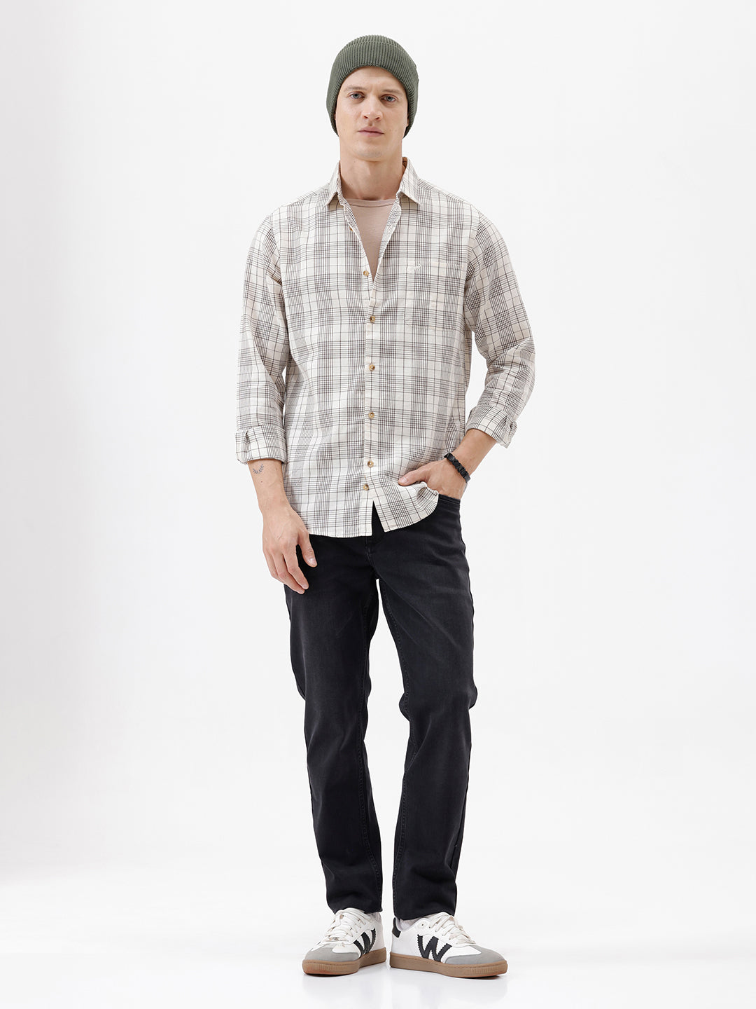 A man wearing a long-sleeve button-up plaid shirt in beige, white, and gray, paired with dark pants, standing against a plain white studio background.