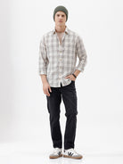 A man wearing a long-sleeve button-up plaid shirt in beige, white, and gray, paired with dark pants, standing against a plain white studio background.
