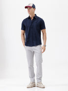 A man wearing a dark navy blue short-sleeve button-down shirt and light gray trousers, standing against a plain white studio background.