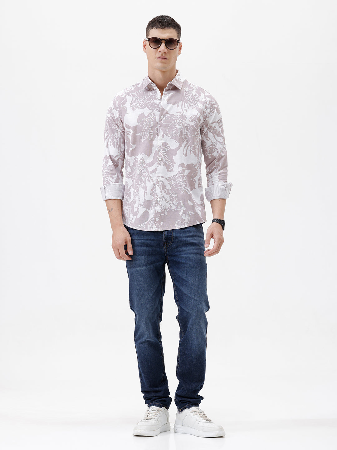 A man wearing a long-sleeve button-up shirt with a light gray and white abstract floral pattern, paired with dark blue jeans, standing against a plain white background.