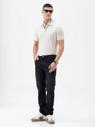 A man wearing a short-sleeve polo shirt in cream with subtle horizontal textured stripes, paired with dark jeans, standing against a plain white studio background.
