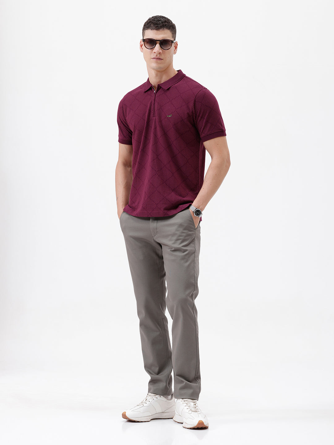 A man wearing a short-sleeve polo shirt in deep burgundy with a subtle textured geometric pattern, styled with gray trousers, sunglasses, and a watch against a plain white background.