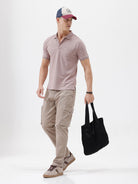 A man modeling a short-sleeve polo shirt in a dusty mauve-pink color, paired with khaki cargo pants and a baseball cap, against a plain light gray background.