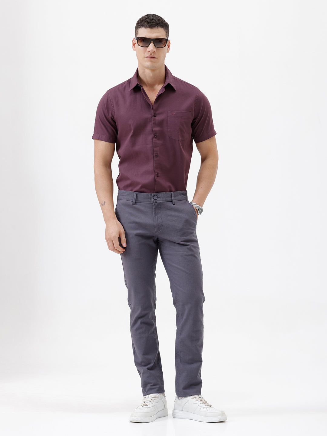 A person wearing slim-fit charcoal gray chino pants, paired with a dark purple shirt and white sneakers, standing against a plain white studio background.