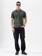 A man wearing a dark olive green short-sleeve polo shirt with a chest pocket, paired with dark jeans, standing against a plain light gray background.