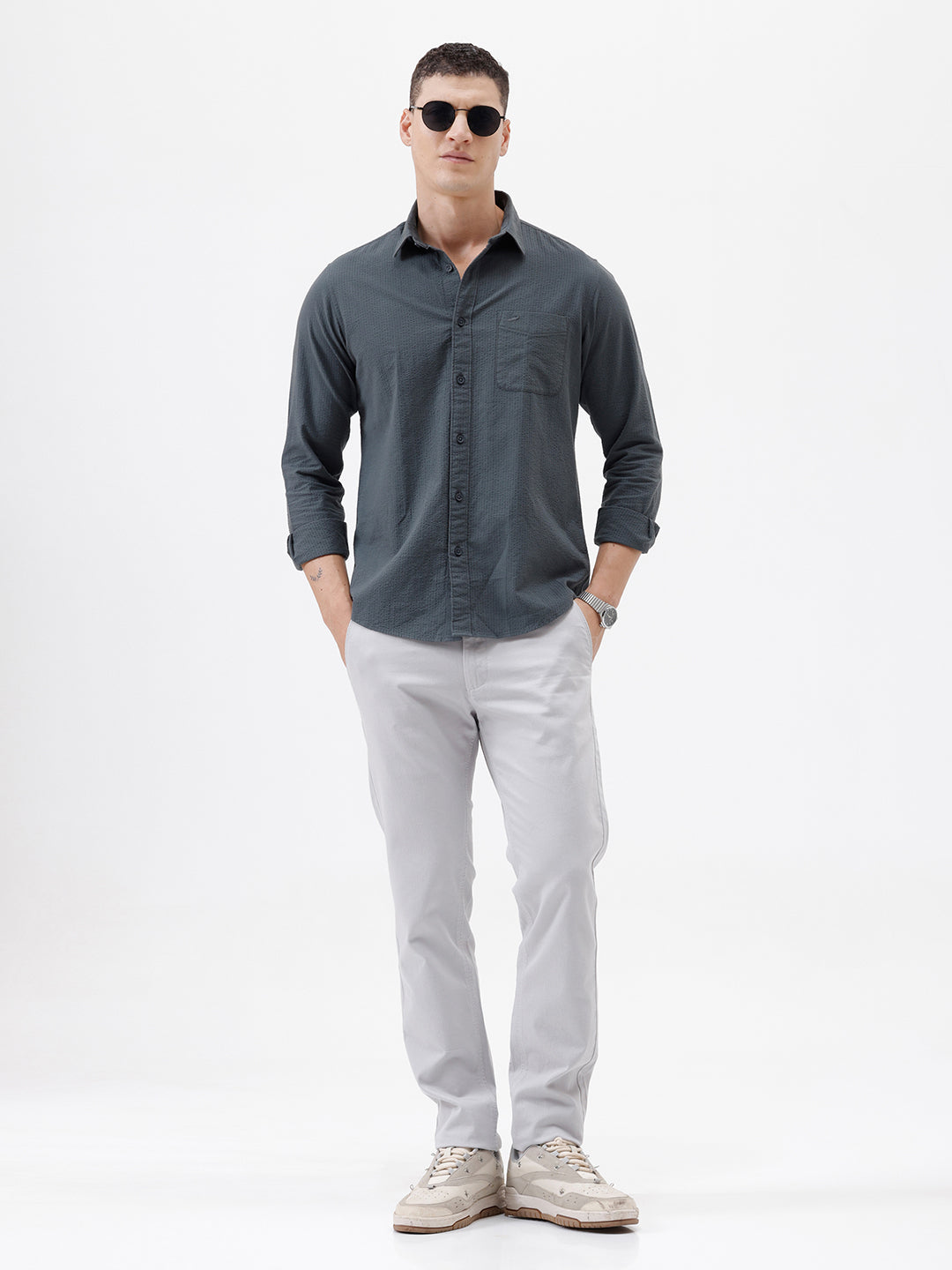 A man wearing a dark charcoal gray long-sleeve button-down shirt with a chest pocket, paired with light gray trousers, standing against a plain white studio background.
