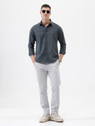 A man wearing a dark charcoal gray long-sleeve button-down shirt with a chest pocket, paired with light gray trousers, standing against a plain white studio background.