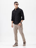 A man wearing a black long-sleeve button-up shirt with a subtle tonal floral pattern, paired with beige cargo pants and dark sunglasses, standing against a plain white background.
