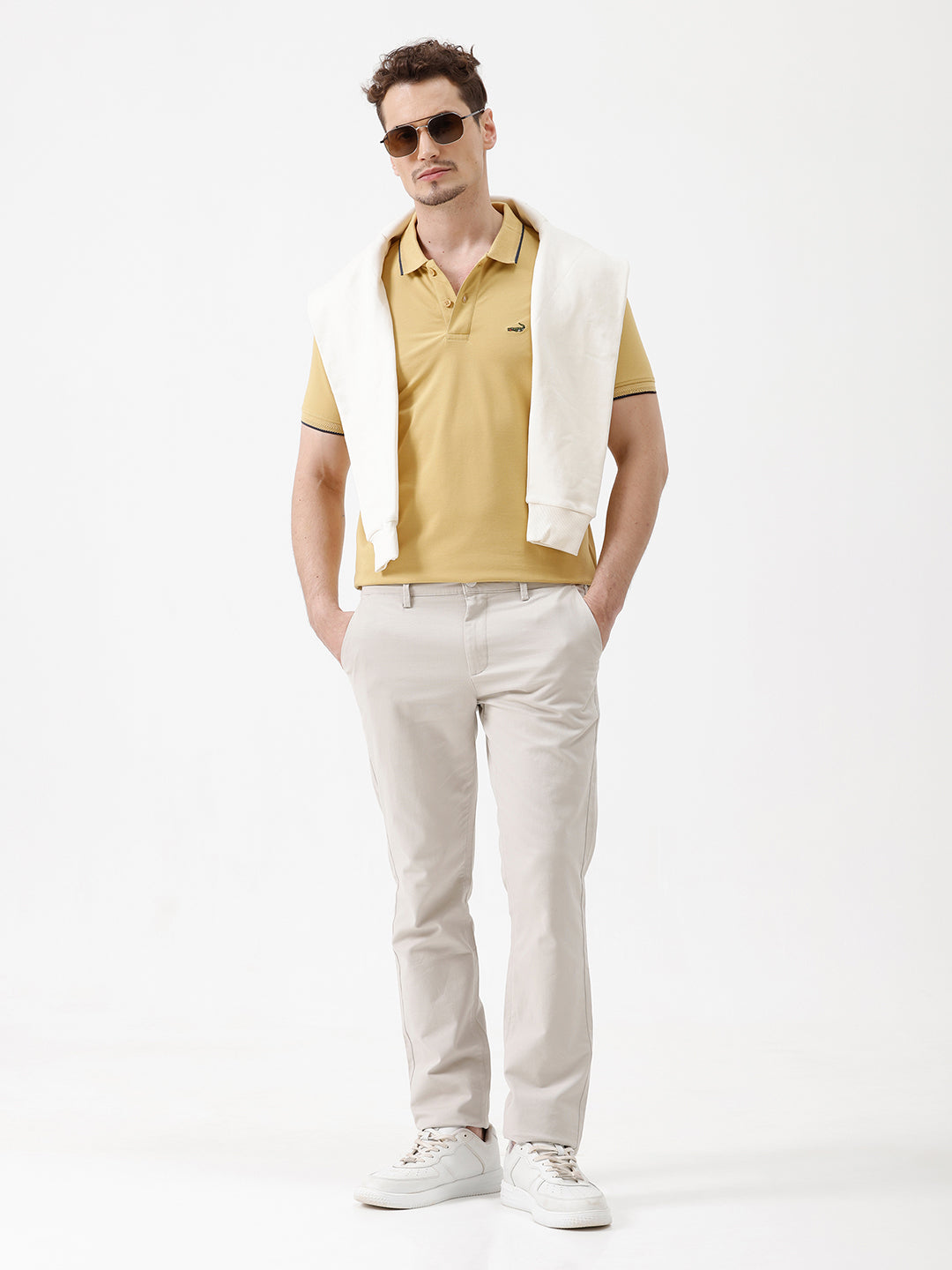 A man wearing a light yellow Lacoste polo shirt with dark piping on the collar and sleeve cuffs, paired with beige trousers, against a plain white studio background.