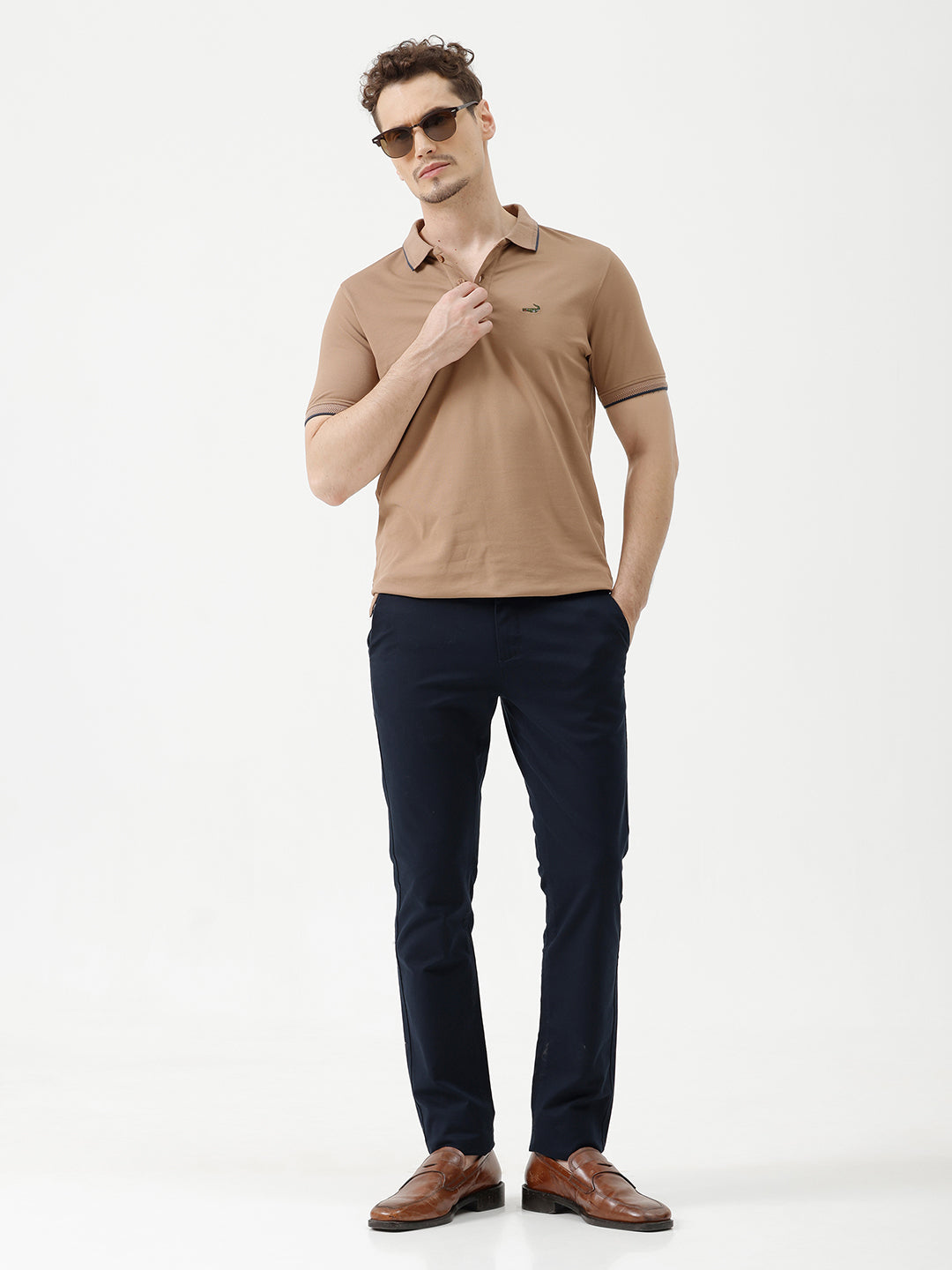 A man wearing a tan short-sleeve polo shirt with dark blue piping on the collar and sleeve cuffs, paired with dark navy pants, against a plain light gray background.