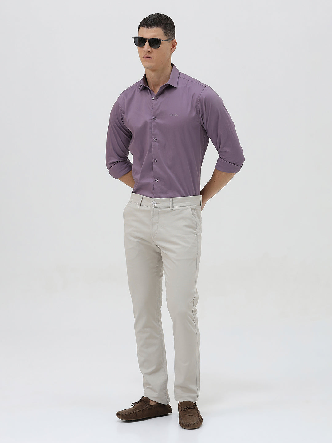 A man wearing a purple long-sleeve button-down shirt with rolled-up sleeves and light beige trousers, standing against a plain light gray background.