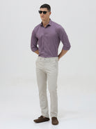 A man wearing a purple long-sleeve button-down shirt with rolled-up sleeves and light beige trousers, standing against a plain light gray background.